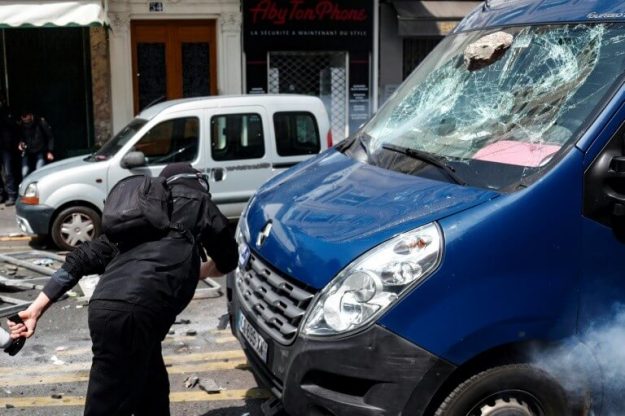 A person dressed in black and hooded launches stones on the windshield of a van, before the start of parades on May 1st in Paris.